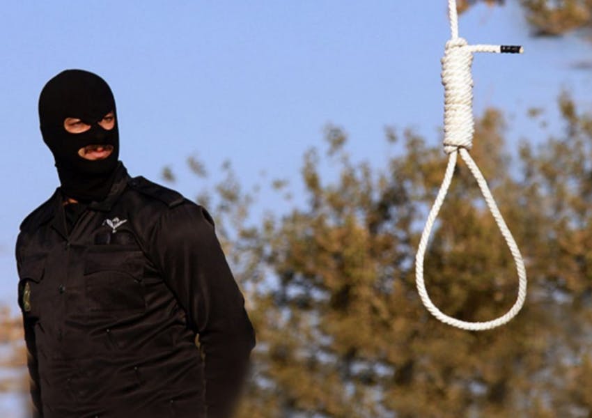 یک مرد با لباس و کلاه نقاب‌دار سیاه در کنار طناب دار دیده می‌شود