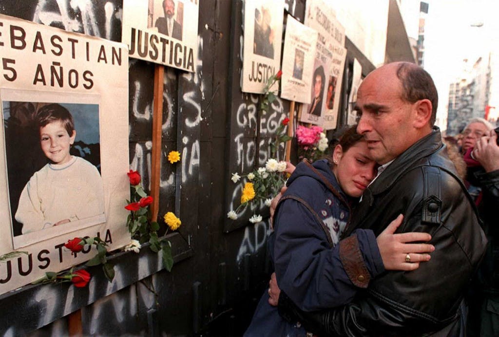تصویر قربانیان بر روی دیوار دیده می‌شود. یک مرد و زن جمقابل دیوار عزاداری می‌کنند