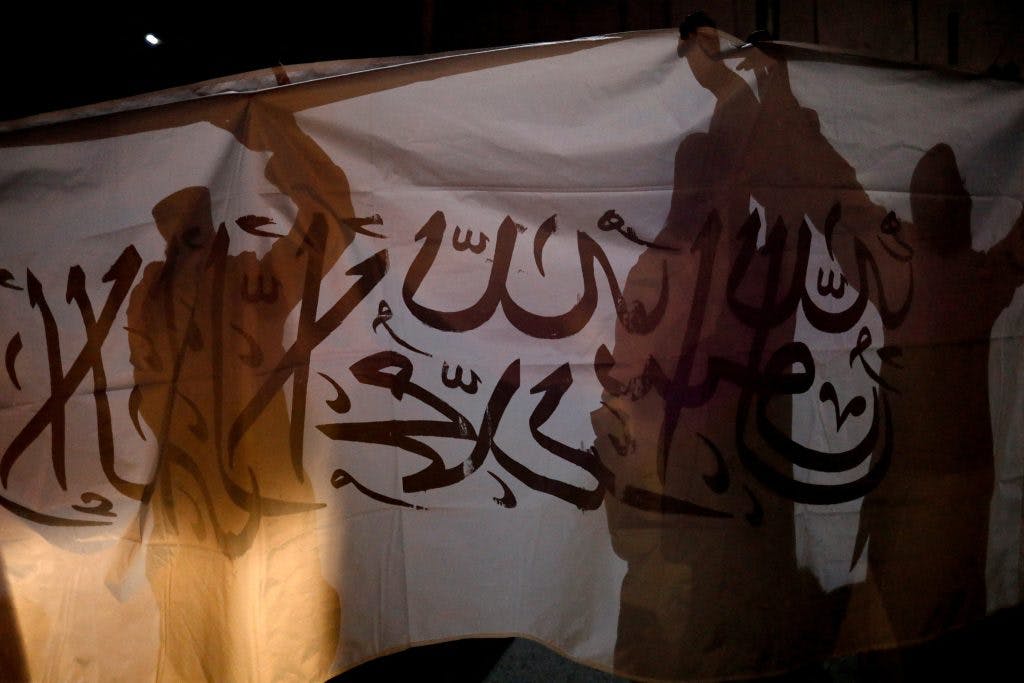پرچم طالبان در پس‌زمینه، و سایه چند طالب سلاح به دست روی آن