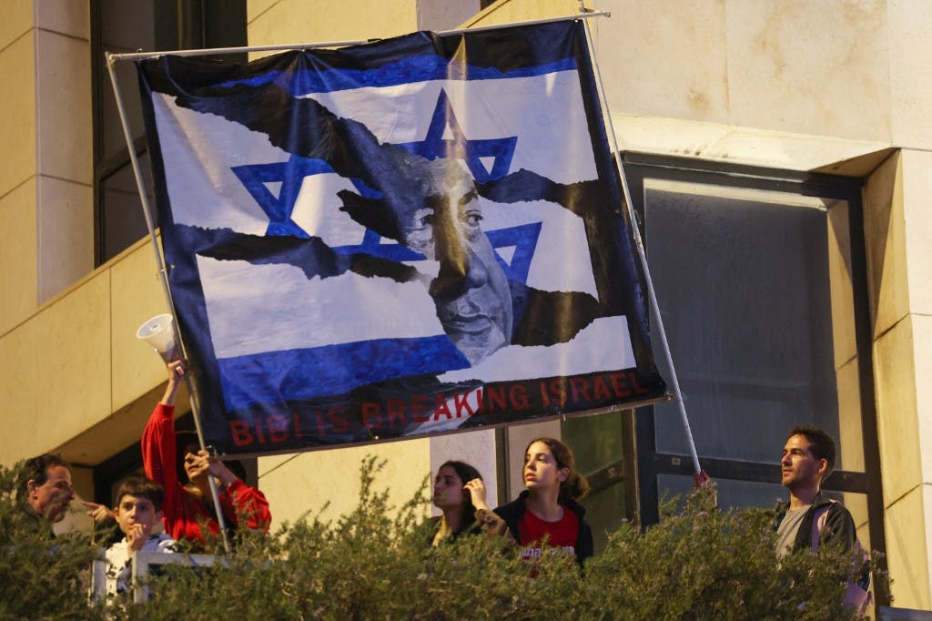 معترضان اسرائیلی بنری را که بنیامین نتانیاهو را در میان پرچم از هم گسیخته اسرائیل نشان می‌دهد، بلند کرده‌اند. تاریخ عکس: ۲۵ مارس ۲۰۲۳ (عکس از احمد قرابلی / خبرگزاری فرانسه)