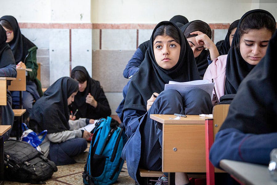 تصویر چند دانش‌آموز دختر را در یک کلاس درس در ایران نشان می‌دهند. چند نفر از آن‌ها روی زمین نشسته‌اند.