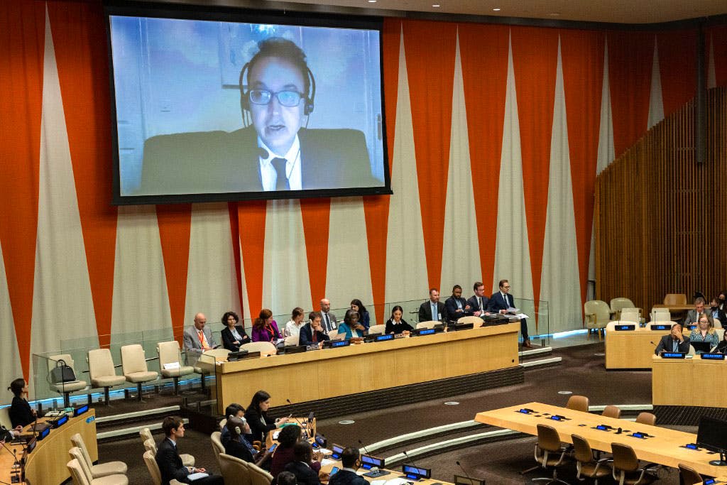 در تصویر، نمای عمومی از سالن مقر سازمان ملل در نیویورک و صفحه بزرگ ویدئو بر روی دیوار دیده می‌شود که در آن جاوید رحمان، گزارشگر ویژه حقوق بشر در ایران، در حال سخنرانی است.