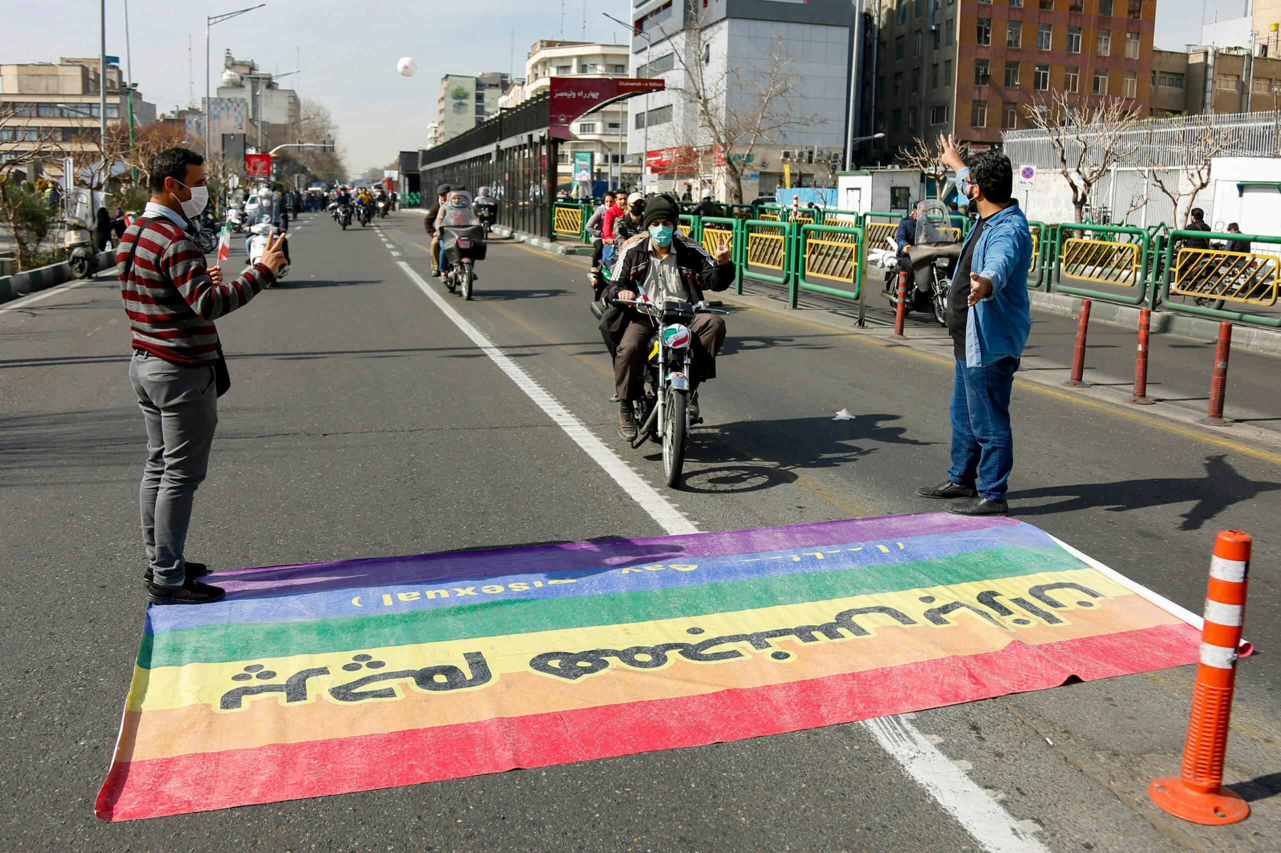 تصویر: در یک راهپیمایی در ایران مردم دعوت به عبور از پرچم رنگین‌کمانی می‌شوند درحالی‌که به فحش بر روی پرچم نوشته شده «پرچم همجنس‌بازان»