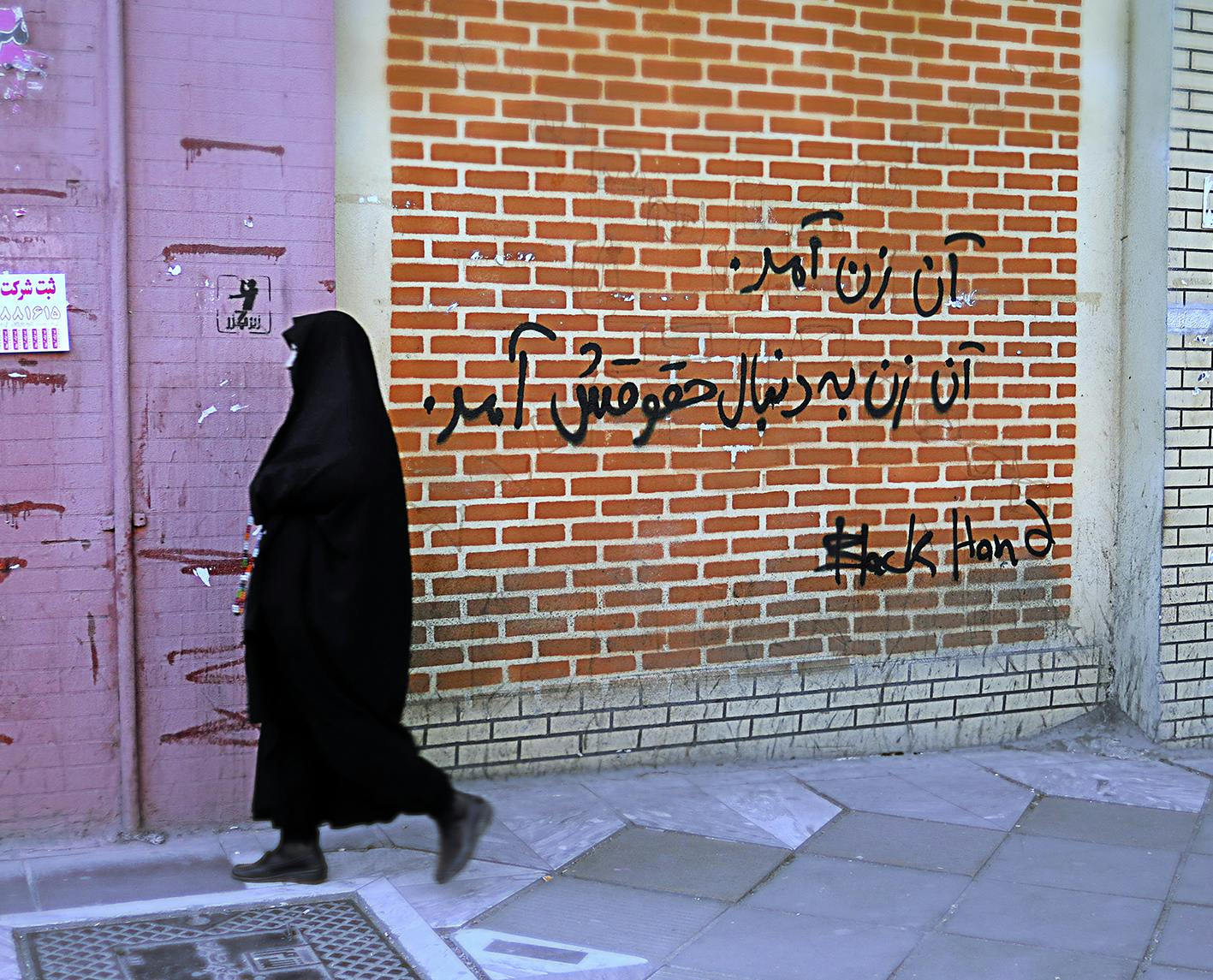 تصویری از یک گرافیتی (دیوارنگاری) با عنوان «آن زن آمد، آن زن به‌دنبال حقوقش آمد». یک زن با حجاب چادر از جلوی این دیوار در حال گذشتن است.