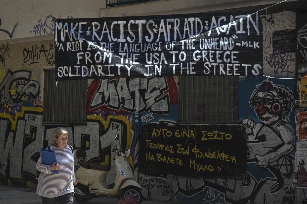 زنی در حال عبور از یکی از کوچه‌های محله اگزارخیا آتن. بر دیوار یک پلاکارد به چشم می‌خورد که بر روی آن نوشته شده است از آمریکا تا یونان همبستگی در خیابان. عکس از AFP