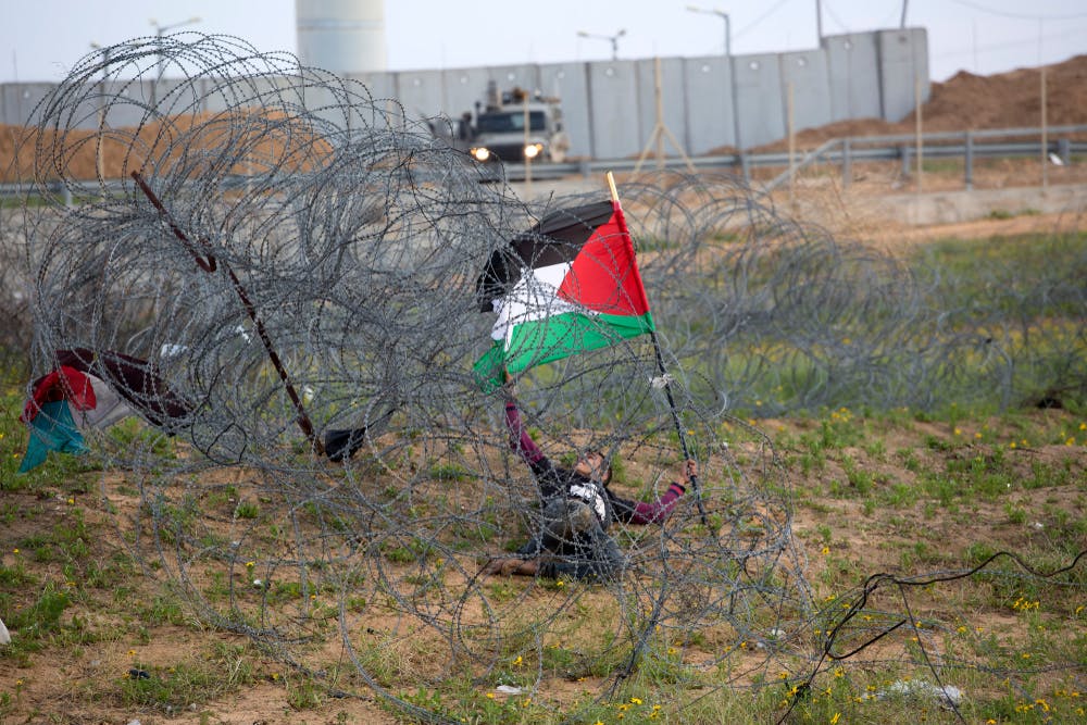 کودک فلسطینی با پرچم فلسطین در زیر سیم‌های خاردار گیرافتاده. سربازان اسرائیلی از دور نظاره‌گرند