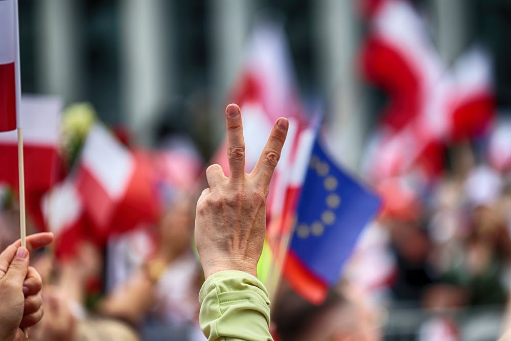 انگشت یک فرد با نشانه پیروزی در پس‌زمینه پرچمهای لهستان و اتحادیه اروپا