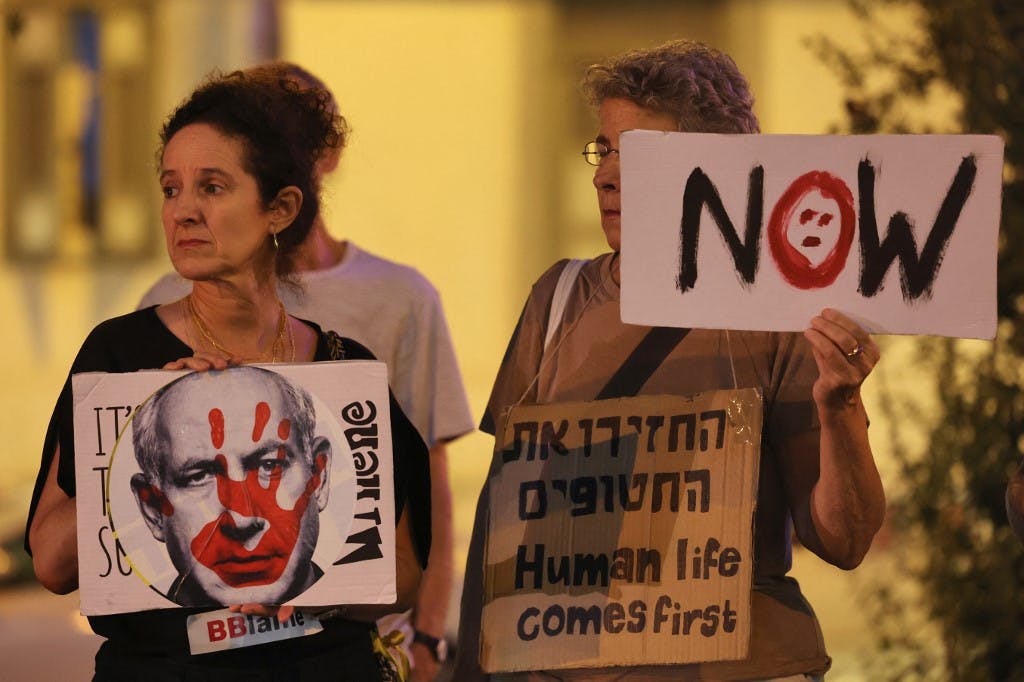تظاهرات در مخالفت با جنگ در تل‌آویو. دو زن کاغذهایی را بر دست دارند. بر روی یکی از کاغذ نوشته ها تصویر نتانیاهو که یک دست خونی بر صورت او دیده می‌شود. و دیگری هم به انگلیس نوشته شده نه.