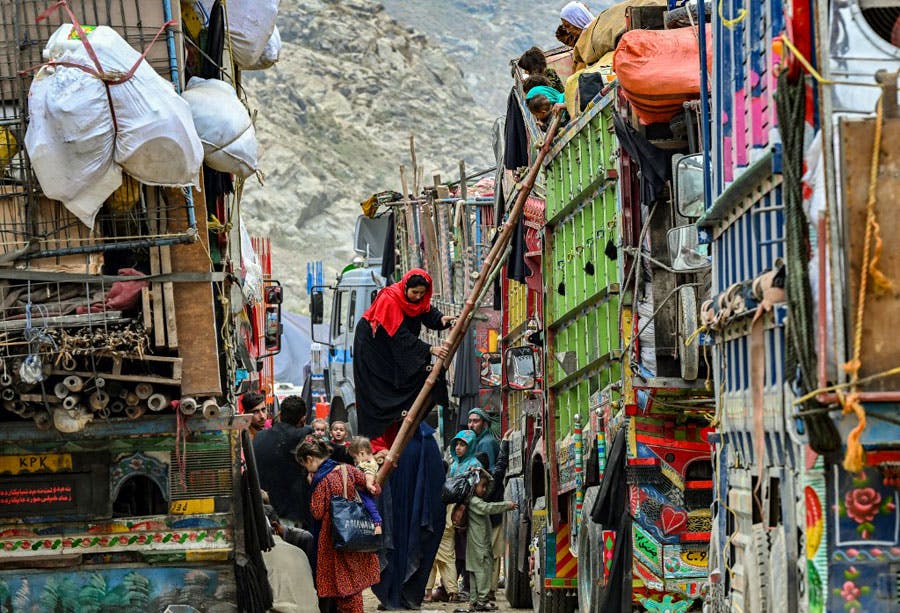 زنان و مردان افغان در یک مرکز نگهداری مهاجران در پاکستان در میان دو ردیف کامیون‌ در حال بار کردن کامیون‌ها با نردبام هستند تا به کشورشان بازگردند. در پس‌زمینه تصویر کوهستان دیده می‌شود.