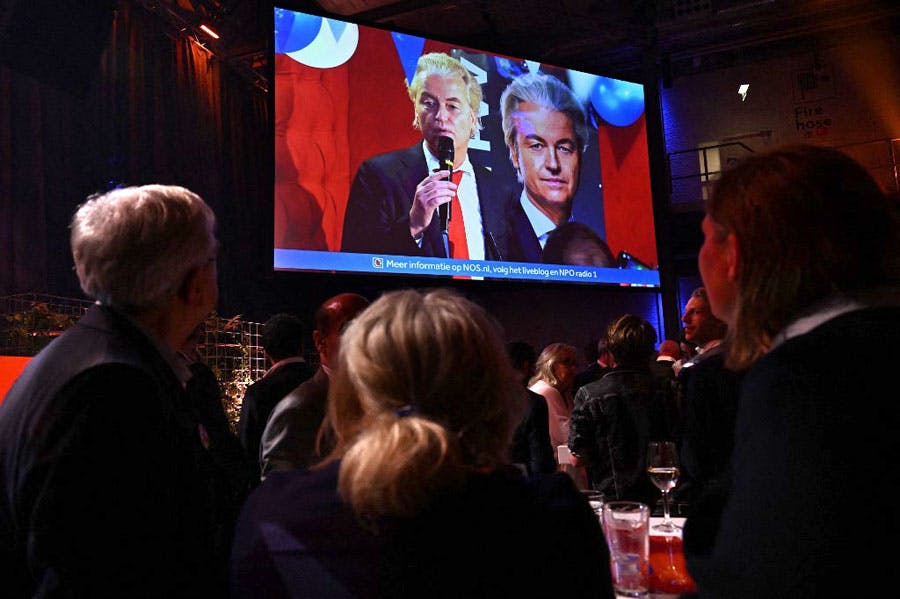 در تصویر، بر زمینه‌ای تیره اعضای حزب «برای آزادی» (VVD) دیده می‌شوند که به اکران تصویر خیرت ویلدرز رهبر این حزب نگاه می‌کنند. خیرت ویلدرز در کنار تصویر انتخاباتی خود ایستاده و در حال سخنرانی است.