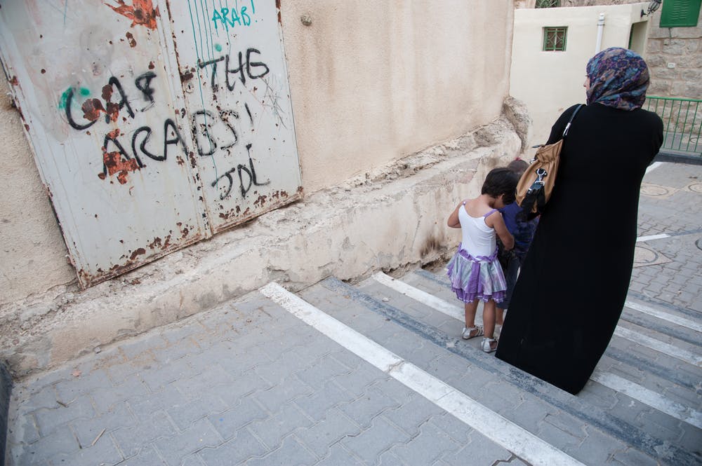 یک زن و کودکانش در الخلیل، کرانه باختری اشغالی از مقابل دیواری که روی آن با اسپری نوشته شده «عرب‌ها را با گاز بکشید» رد می‌شوند. تاریخ عکس ۲۲ اکتبر ۲۰۱۲ - عکس از شاتراستاک