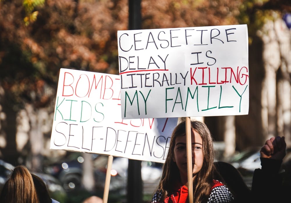کودک بنری به زبان انگلیسی در دست دارد که با رنگ‌های سبز و قرمز و سیاه روی آن نوشته: تأخیر در آتش‌بس واقعاً دارد خانواده‌ام را می‌کشد. بنر دیگری نیز پشت آن هست که رویش نوشته: بمباران‌کردن کودکان دفاع از خود نیست.
