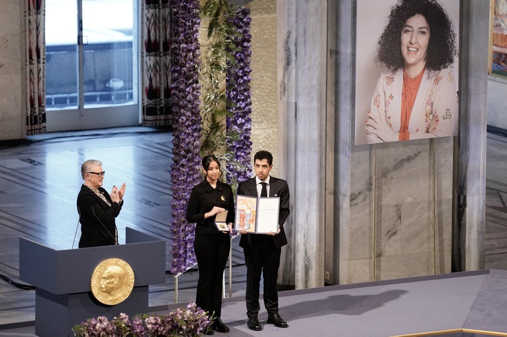 رئیس کمیته نوبل در حال دست زدن، و فرزندان نرگس محمدی در سمت راست تصویر روی سن جایزه را در دست دارند. روی دیوار تصویر بزرگ نرگس محمدی است.