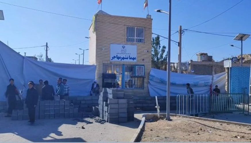 یکی از دروهای ورودی مهانشهر _ اردوگاه مهاجران افغانستانی در میبد را با بلوک سیمانی مسدود می‌کنند. تابلویی که بر آن نوشته شده »مهمانشهر مهاجر« بر روی دیوار دیده می‌شود.