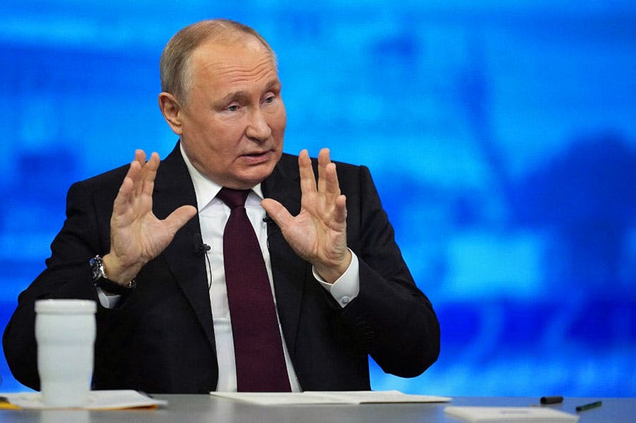 ولادیمیر پوتین، رئیس جمهوری روسیه در کنفرانس رسانه‌ای پایان سال خود در مسکو پشت یک میز در حال صحبت‌کردن است. او دو دست خود را بالا برده و موضوعی را دارد توضیح می‌دهد. پس‌زمینه‌ی او آبی‌رنگ است.