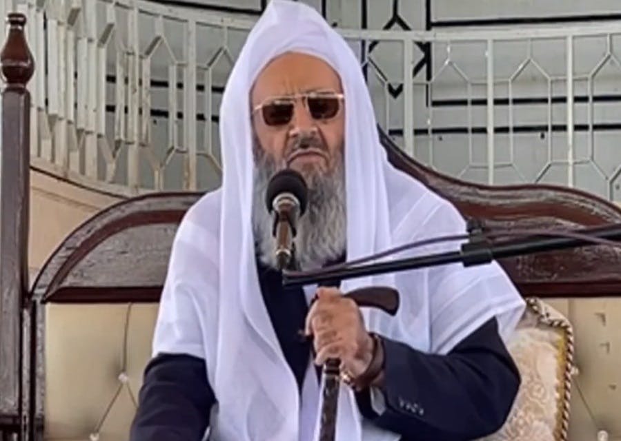 عبدالحمید اسماعیل‌زهی با ردای سفید و عینک آفتابی و عصا در دست پشت بلندگو