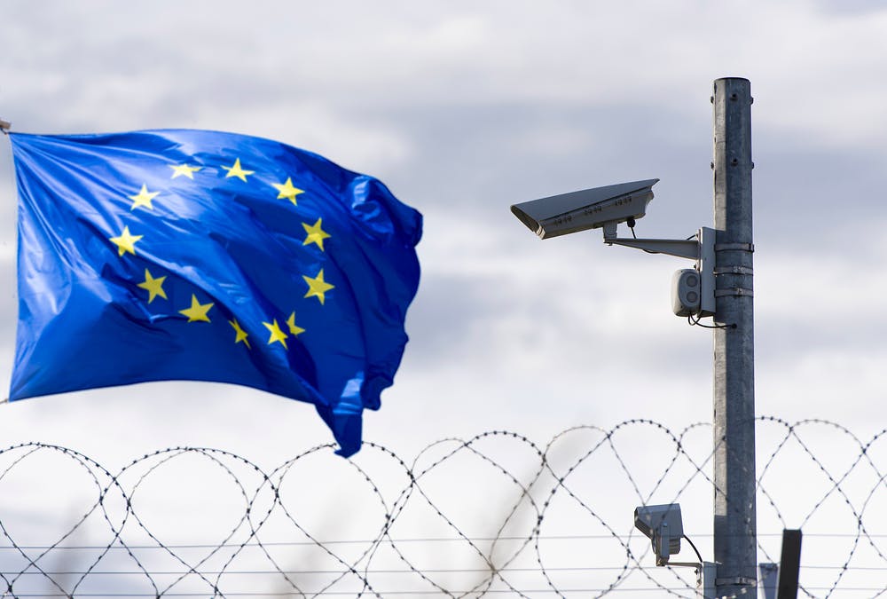 در سمت چپ عکس پرچم اتحادیه اروپا (آبی‌رنگ با دایره ستاره در مرکز)، سمت راست یک دوربین مداربسته و پایین عکس سیمهای خاردار
