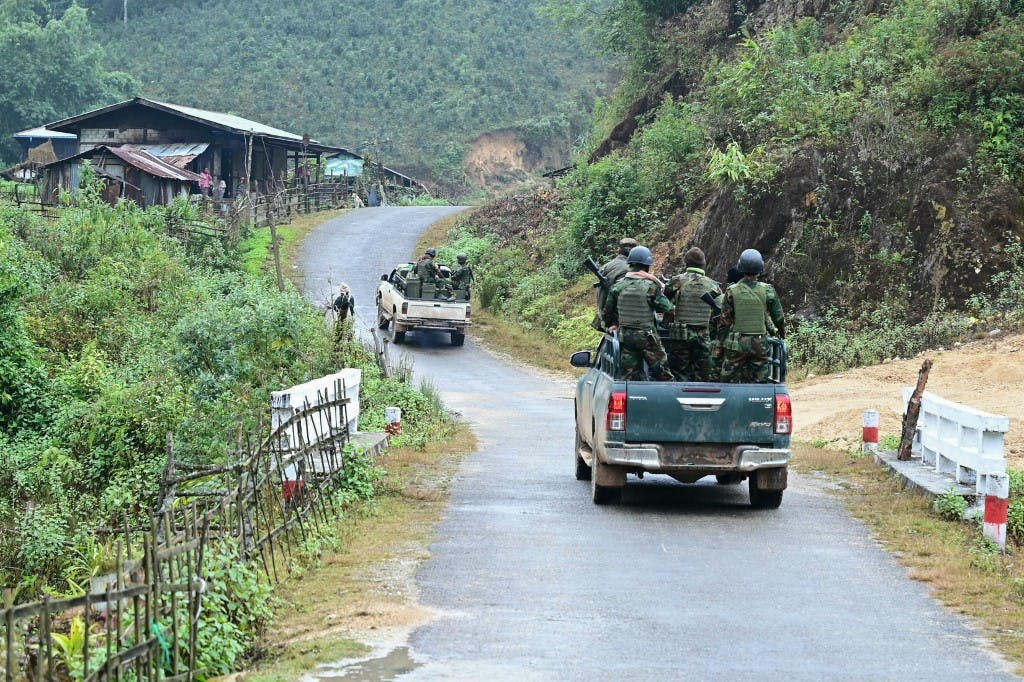 تصویر از پشت دو کامیون پر از سرباز را نشان می‌دهد که در حال حرکت از جاده‌ای باریک از میان جنگل‌های انبوه میانمار در آسیای جنوب شرقی هستند