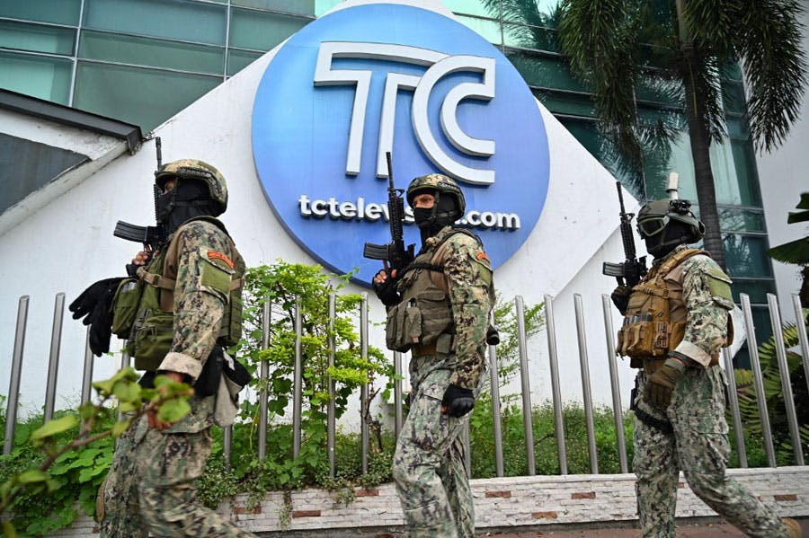 سه سرباز اکوادوری در مقابل ساختمان شبکه تلویزیونی تی‌سی سلاح به‌دست با چهره‌های پوشیده در حال حرکت‌اند. در پسزمینه تصویر روی ساختمان لوگوی شبکه تی‌سی دیده می‌شود.