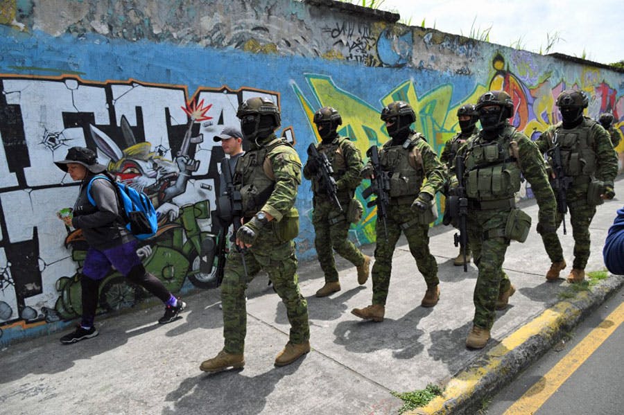 شش سرباز ارتش اکوادور با سلاح‌های خود در پیاده‌روی خیابانی از کنار یک دیوار با نقش‌های گرافیتی و یک زن و یک جوان عبور می‌کنند.
