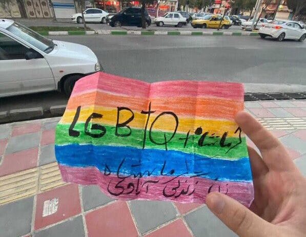 عکس تزئینی از فضای مجازی، یک دست را نشان می‌دهد که در یک خیابان داخل ایران، کاغذی رو به دوربین گرفته که بر آن پرچم رنگین‌کمان کشیده شده و نوشته شده ال‌جی‌بی‌تی‌کیو پلاس کرمانشاه