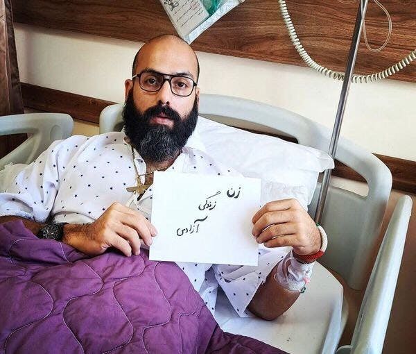 تصویری از خالد پیرزاده، زندانی سیاسی روی تخت بیمارستان. او کاغذی را که روی آن نوشته شده «زن زندگی آزادی» در دست دارد.