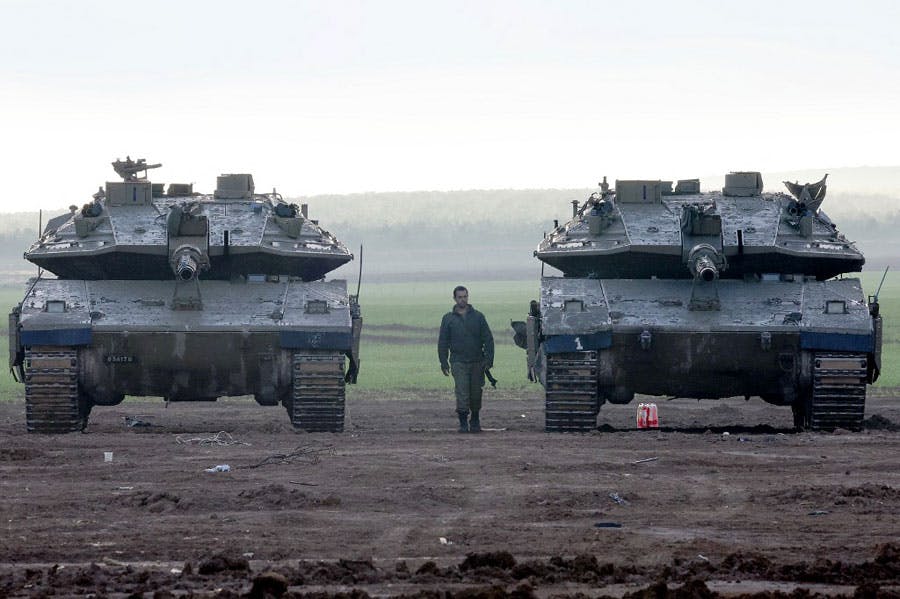 در این تصویر یک سرباز اسرائیلی در میان دو تانک ارتش این کشور در غزه دیده می‌شود. هوا مه‌آلود است و در پسزمینه دشت سبزی قرار دارد. زیر یکی از تانک‌ها نیز یک گالن دیده می‌شود.