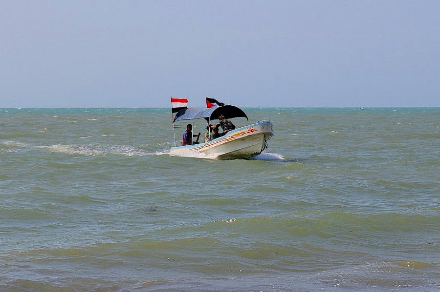در تصویر قایق کوچکی با چهار سرنشین بر روی آب دیده می‌شود. پرچم‌های یمن و فلسطین روی قایق در باد تکان می‌خورند.