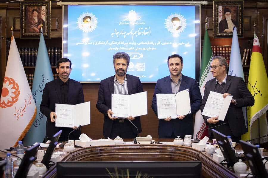 چهار مرد که هر کدام نسخه‌ای از یک قرارداد را در دست دارند، جلوی صفحه نمایش آبی‌رنگی با عکس خامنه‌ای و خمینی ایستاده‌اند و رو به دوربین لبخند میزنند.
