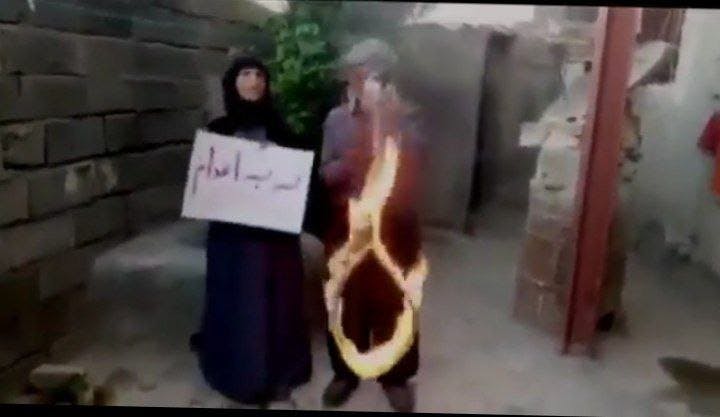 زن و مردی در حیاط خانه‌ای نیمه‌ساخته زن نوشته‌ی نه به اعدام در دست دارد و مرد ظناب داری را آتش زده است