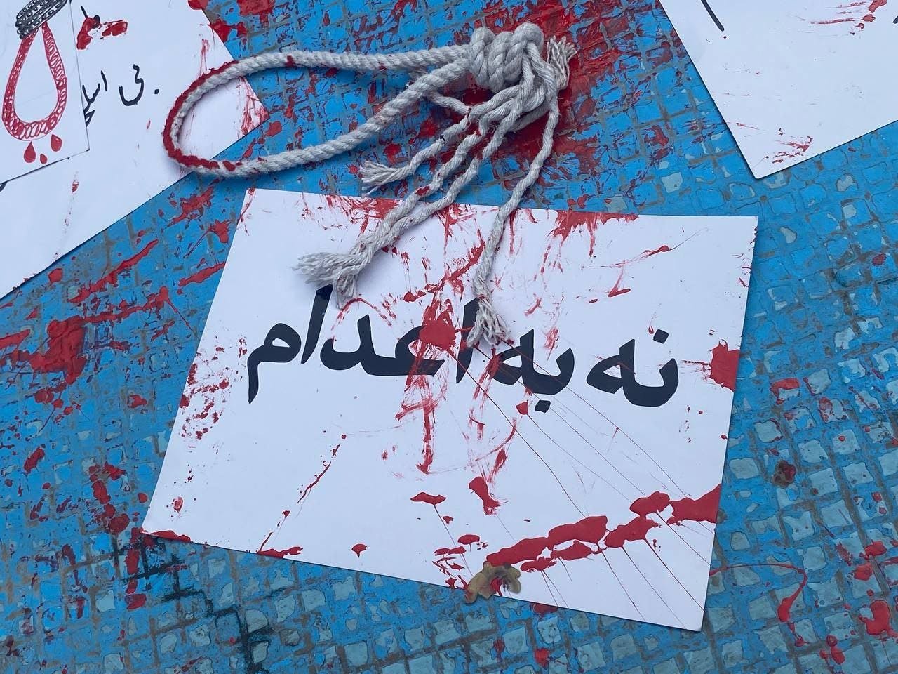روی کاغذ سفیدی نوشته شده است نه به اعدام و روی آن رنگ قرمز پاشیده شده و طنابی پاره روی آن افتاده