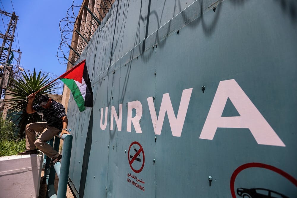 عکس یک دیوار آهنی که روی آن نوشته UNRWA. بالای دیوار سیمهای خاردار است. یک جوان فلسطینی با پرچم فلسطین به دیوار تکیه داده.