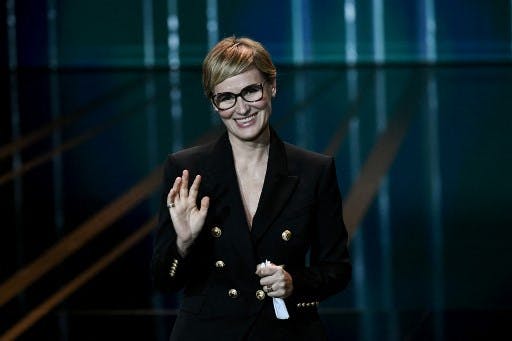 ژودیت گدرش، بازیگر فرانسوی، با موهای بلوند کوتاه و عینک و کت سیاه، دست راستش را بالا گرفته و در دست چپش میکروفون است
