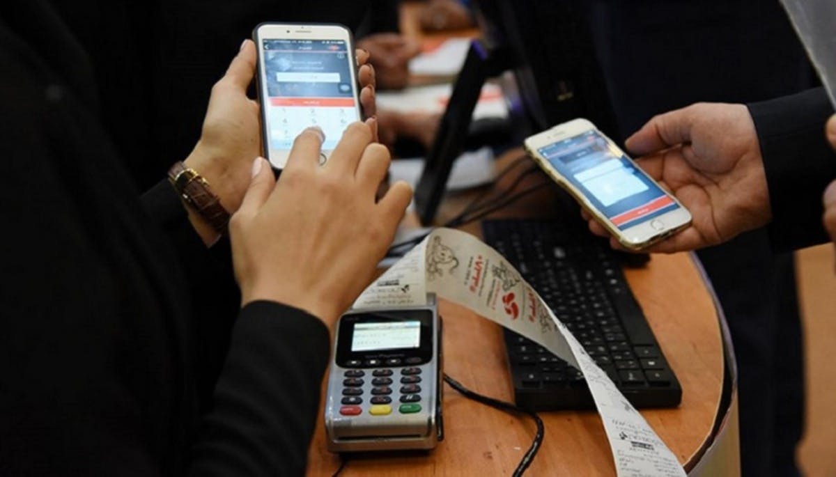 تصویری از دو نفر در حال پرداخت پول با اپلیکیشن در یک فروشگاه - روی میز یک دستگاه کارتخوان با رسید خرید دیده می‌شود.