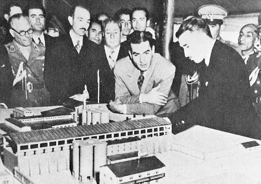 محمدرضا شاه جوان در سال 1955 در حال بازدید از ماکتی مرتبط با طرحهای توسعه‌ای. اطراف او را گروه‌هایی از مقامات گرفته‌اند. محمدرضا شاه با قیافه‌ای کنجکاو به سمت ماکت یک ساختمان خم شده و آن را نگاه می‌کند.