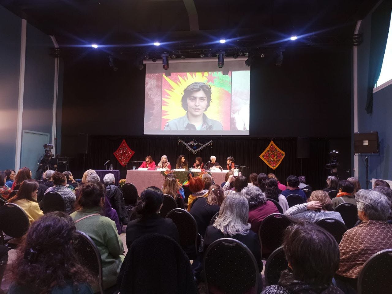تصویر جمعیت حاضر در کنفرانس. روی دیوار صفحه نمایشی هست که عکس یکی از شهدای زن کردستان را نمایش میدهد.