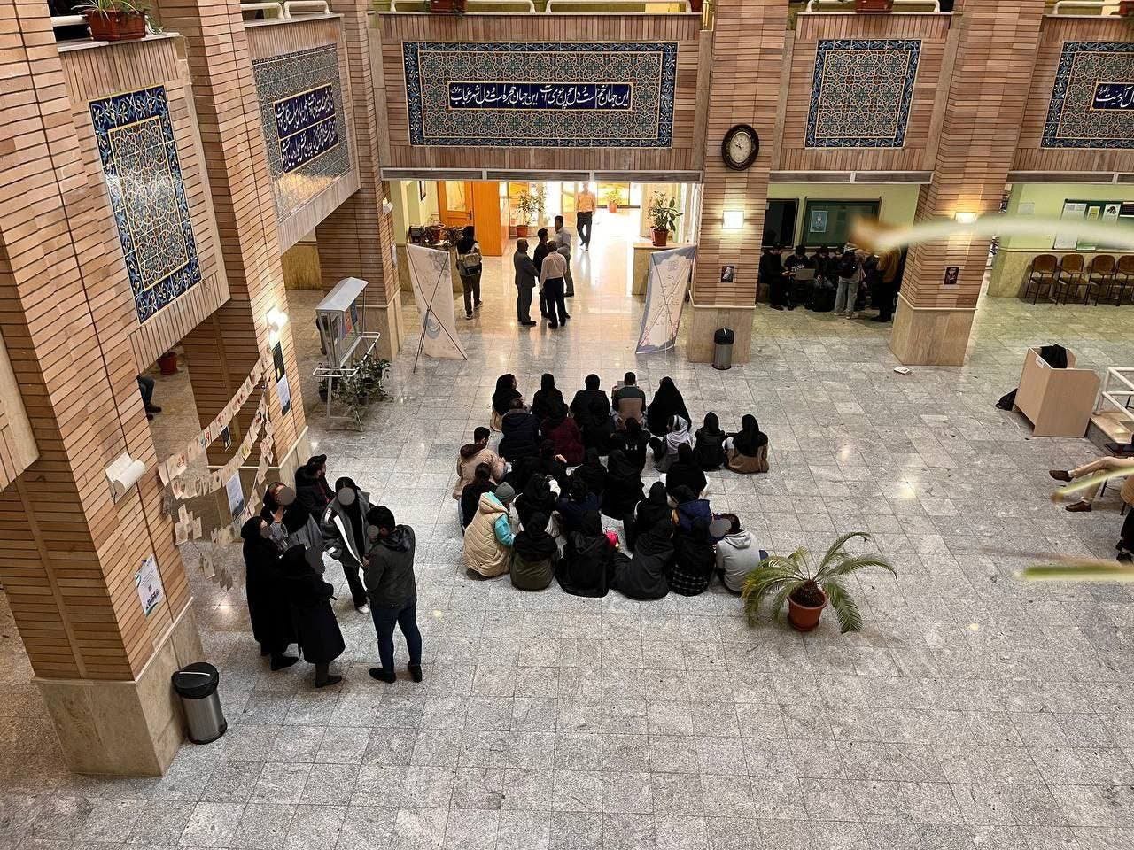 دوربین از بالا طبقه همکف دانشکده روانشناسی دانشگاه بهشتی را نشان می‌دهد که گروهی از دانشجویان در اعتراض به فضای امنیتی دانشگاه کف آن نشسته و تحصن کرده‌اند.