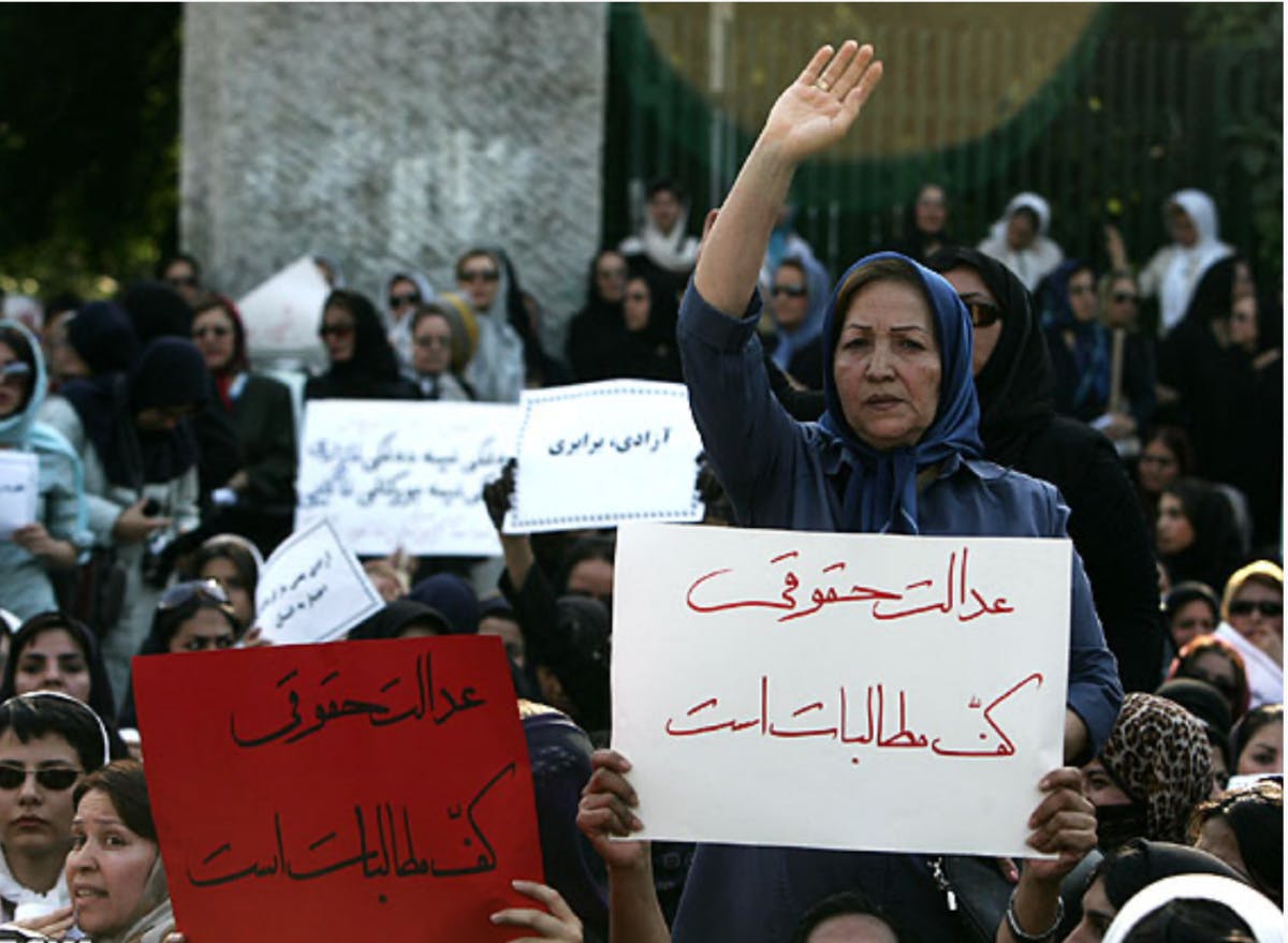 تصویری از یک تجمع زنان. در این تصویر زن میانسالی که دستان خود را بالا گرفته دیده می‌شود. مقابل او زن دیگری یک پلاکارد را بالا برده که روی آن نوشته شده عدالت حقوقی کف مطالبات است.
