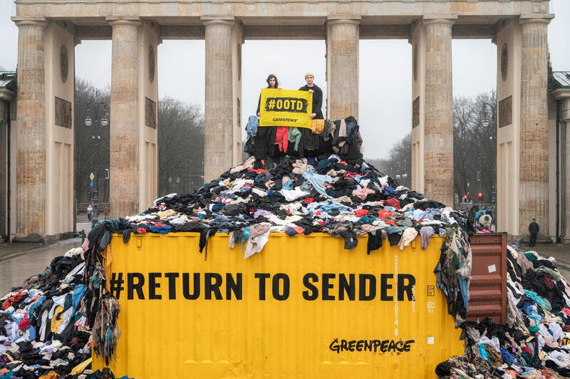 اعتراض فعالان مدنی در هفته مد برلین، پایتخت آلمان را نشان می‌دهد که بر یک تپه لباس ایستاده‌اند که بر روی کانتینر زیر آن نوشته شده: به تولیدکننده برگردانده شود تا یادآور مشکلات زباله صنعت مد باشند.