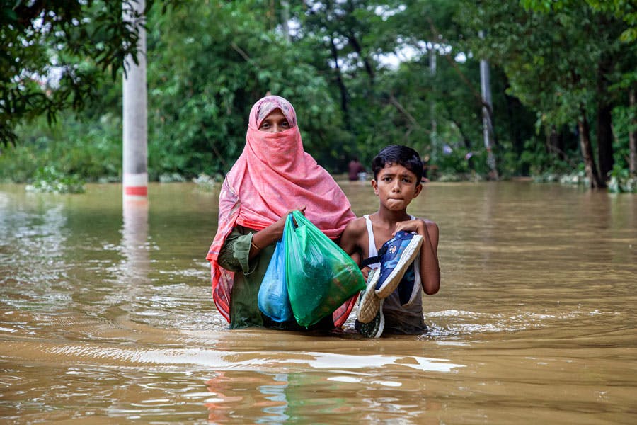 یک زن بنگلادشی و فرزند نوجوانش تا کمر در آب جاری بر اثر سیل ایستاده‌اند و رو به دوربین نگاه می‌کنند. آن‌ها یک جفت کفش و دو کیسه پلاستیکی در دست دارند.