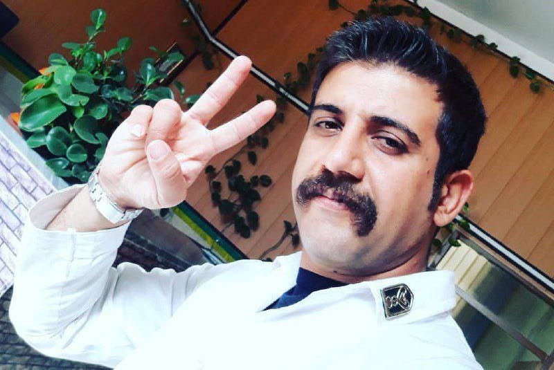 ساسان احمدی، با سبیل پهن و لباس سفید پرستاری، در حالی که دو انگشتش را به نشانه پیروزی بالا آورده و به دوربین نگاه میکند