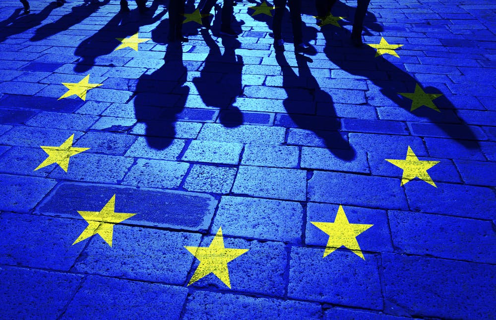 تصویر از سایه چند نفر بر یک سنگ‌فرش که ستاره‌های پرچم اتحادیه اروپا این سایه‌ها را احاطه کرده است.