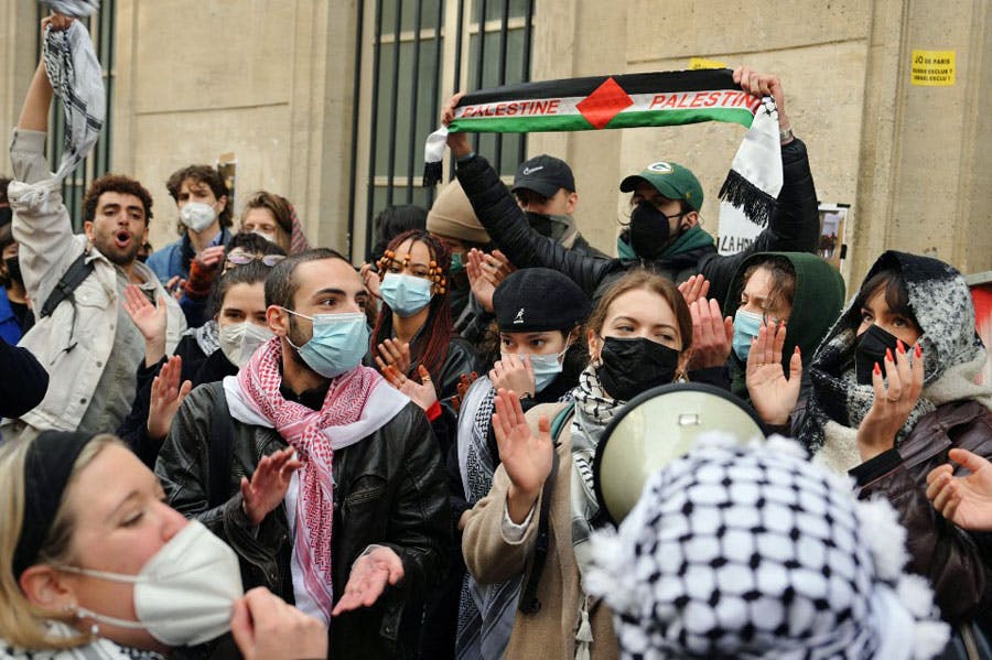 گروهی از دانشجویان که برخی از آن‌ها ماسک پزشکی (متعلق به دوران کرونا) بر چهره دارند پرچم فلسطین را بالا برده‌اند و دست می‌زنند.