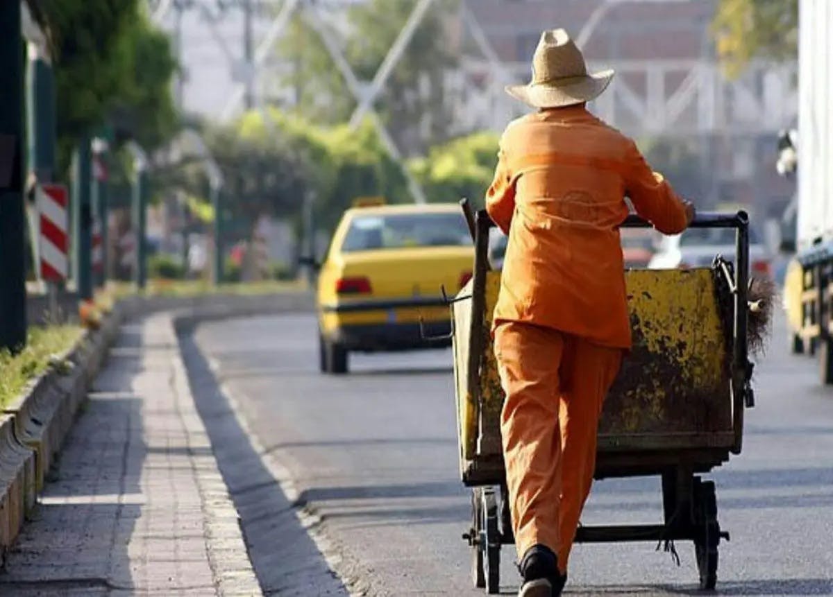 یک کارگر نارنجی‌پوش در حال هُل دادن گاری دستی در خیابان. جلوتر از او یک خودروی زردرنگ تاکسی دیده می‌شود.