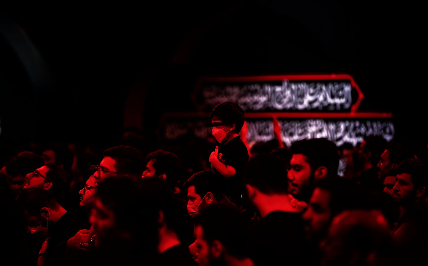 تصویری سیاه و قرمز، از یک هیئت امام حسین. تصویر از زاویه کنار، عزاداران را نشان میدهد و وسط تصویر یک کودک روی دوش مردی دیگر در حال سینه‌زدن است