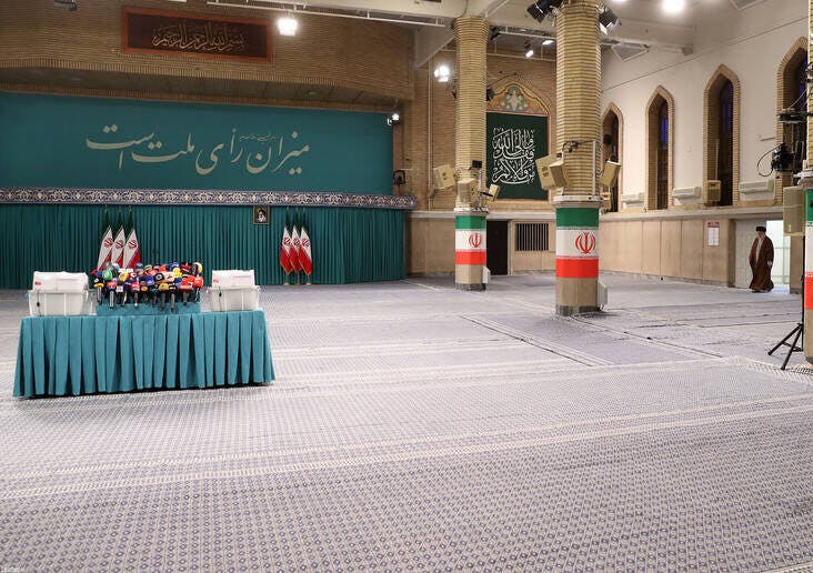 تصویر صحن حسینیه امام خمینی را نشان می‌دهد. در دورترین نقطه سمت راست، علی خامنه‌ای بسیار ریز نشان داده شده که دارد از دری وارد صحن می‌شود. در سمت چپ تصویر، میزی که روی آن گل و