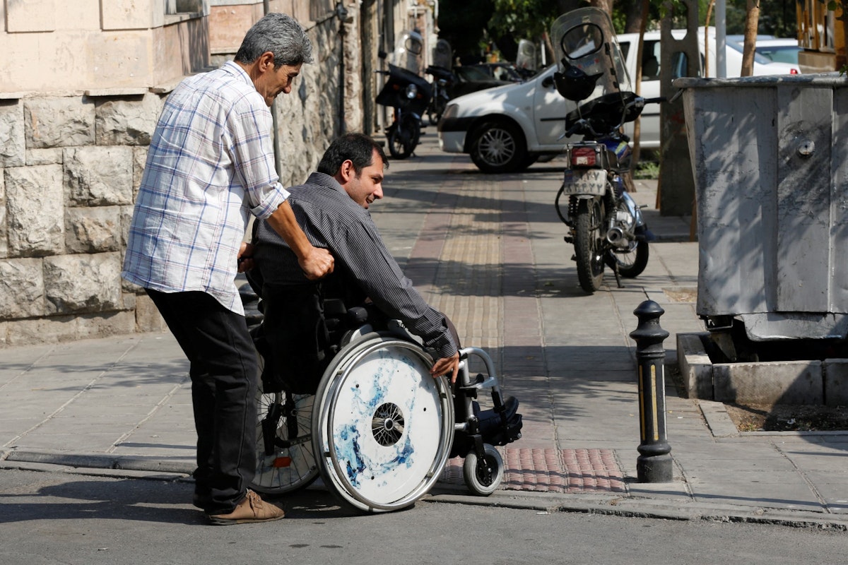 تصویر تزئینی از خبرگزاری فرانسه تلاش برای عبور ازیک پیاده‌رو توسط یک فرد نشسته بر ویلچر و یک نفر که همراه است نشان می‌دهد.