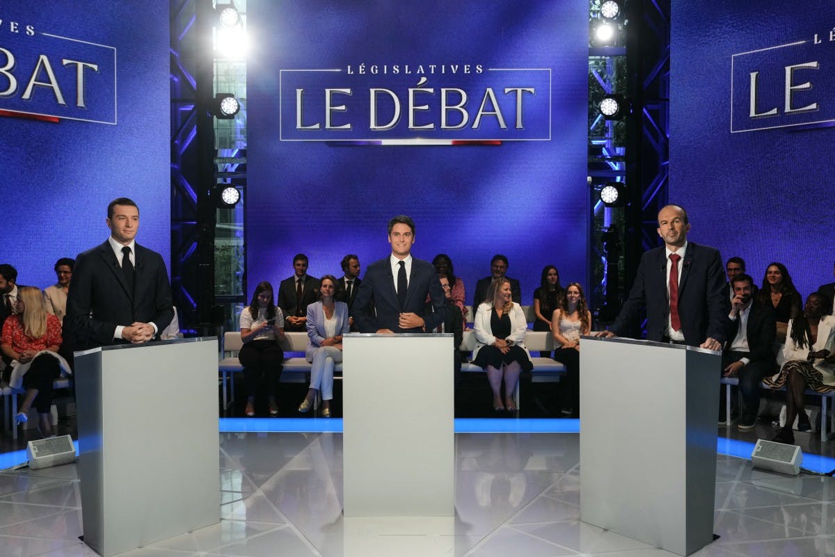 صحنه مناظره تلویزیونی انتخابات مجلس فرانسه، پس‌زمینه صحنه آبی پررنگ است و تماشاچیان نشسته‌اند، در جلوی صحنه سه نامزد مرد پشت میز با فاصله کنار هم ایستاده‌اند