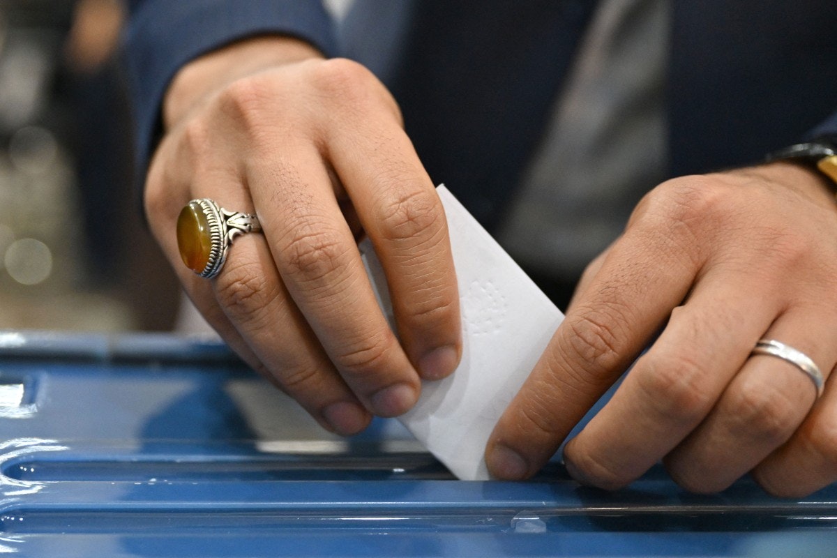 تصویر دو دست که روی دست راست انگشتر عقیق و روی دست چپ انگشتر ساده دیده می‌شود، در حال انداختن برگ رای به صندوق