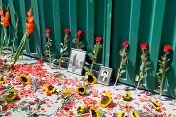 در تصویر گل‌های سرخ و آفتابگردان به همراه عکس‌های اعدام‌شدگان دهه ۶۰ و تابستان ۶۷ پای دروازه بسته سبزرنگ گورستان خاوران نشان داده شده است.